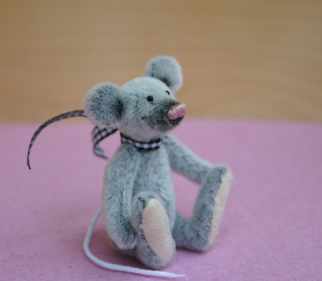 sehr kleine graue Maus mit rosa Nase, dunklem Schnäuzer aus kurzem Flor sitzt auf einem rosa Wollfilz