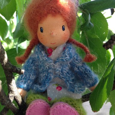 Miniatur Püppchen mit roten Haaren und Sommersprossen sitzt auf einem Ast mit grünen Blättern drum herum