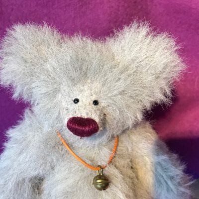 Nahaufnahme eines Waldzwerge Teddys aus hellgrauem Alpaka mit dunkel brombeerfarbener Nase und Glöckchen um den Hals vor dunkel pinken Hintergrund
